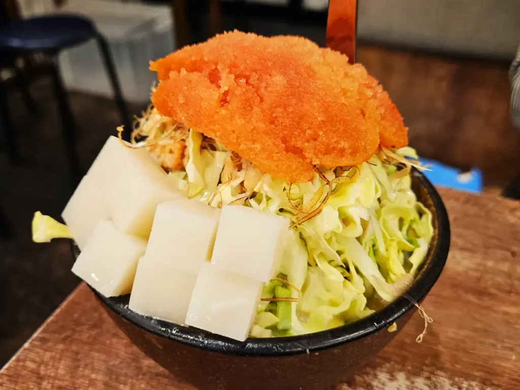 A bowl piled high with raw monjayaki ingredients, featuring shredded cabbage topped with white mochi cubes and a large portion of bright orange spicy mentaiko roe.