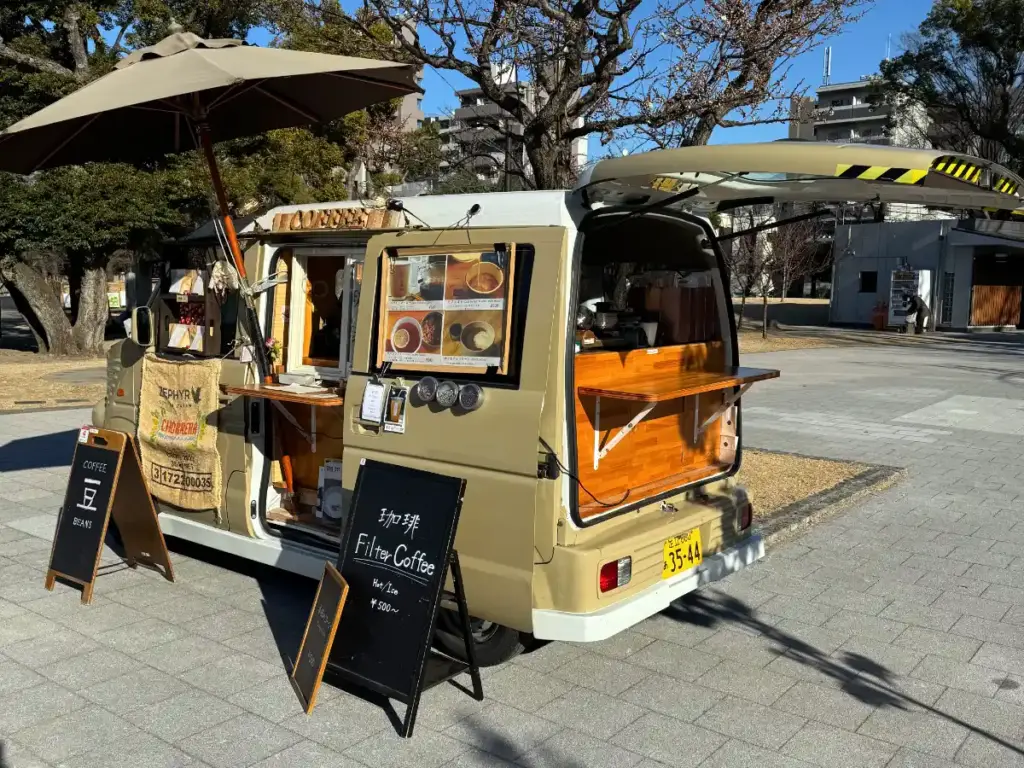A cute, retro beige van converted into a mobile coffee shop parked at Sumida Park Breeze Plaza in Tokyo. The van has a wooden serving counter, a large sun umbrella, and a black board sign offering hot and iced filter coffee.