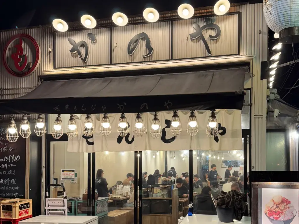 The exterior of Monja Kanoya restaurant on Hoppy Street in Asakusa at night, with a white noren curtain and patrons visible inside.