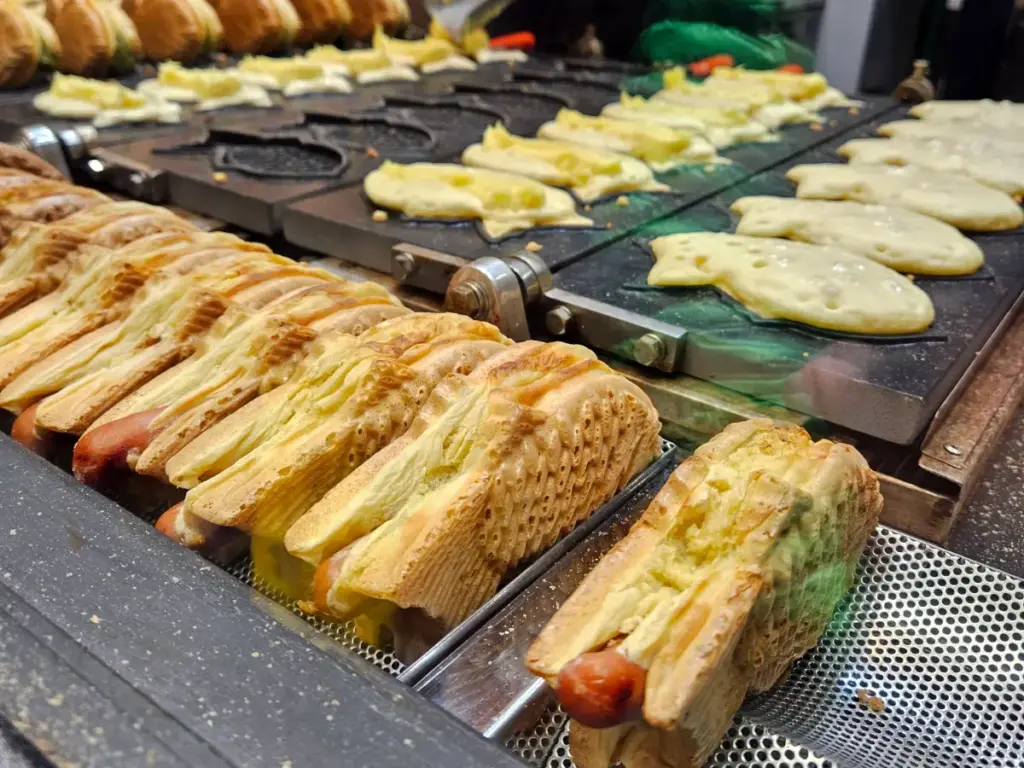 Several fish-shaped taiyaki cooking on a hot iron griddle at a Tokyo street food stall, filled with hotdogs instead of traditional sweet paste.