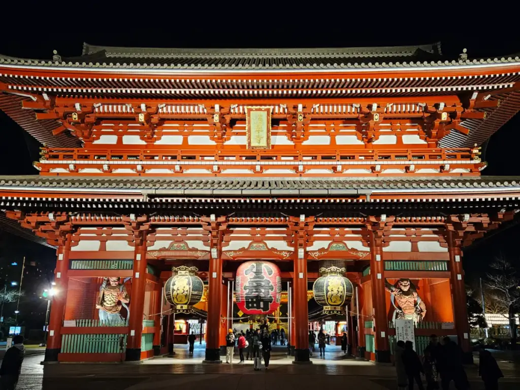 The Hozomon Gate at Senso-ji Temple at night, beautifully illuminated with bright red and white lights. A massive red chochin lantern with black kanji hangs in the center, flanked by Nio guardian statues and traditional architecture.