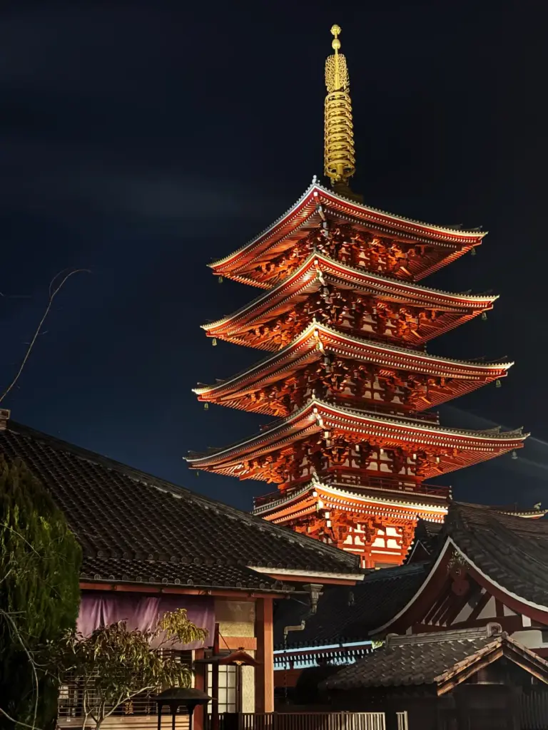 The illuminated five-story pagoda at Senso-ji Temple in Asakusa glows bright red against a dark blue night sky in Tokyo. The view is framed by traditional dark tile roofs and green foliage.