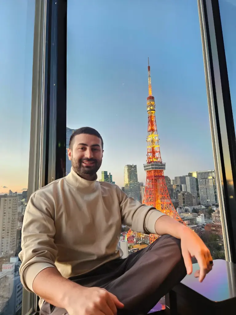 Tim sitting inside the Sky Lounge at The Prince Park Tower Tokyo, casually smiling next to a large floor-to-ceiling window with a view of the Tokyo Tower.