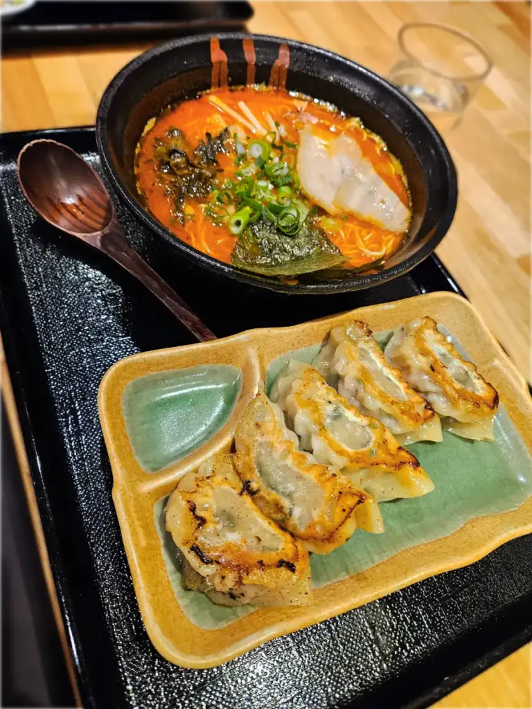 A black tray holding a bowl of hot, spicy red tonkotsu ramen with a wooden spoon, sitting next to a green ceramic plate with pan-fried gyoza dumplings.