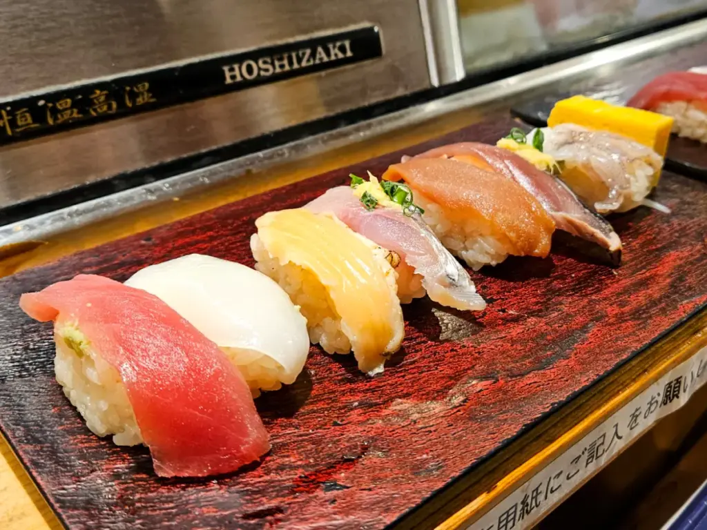 A colorful, fresh assortment of nigiri sushi, including tuna, squid, and salmon, served on a red and black wooden block at a standing sushi counter.