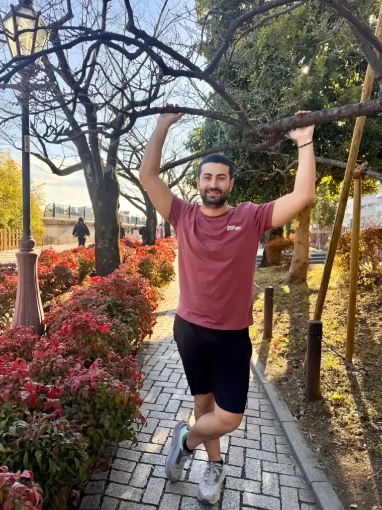 Tim smiles and stretches by holding a bare tree branch during a sunny morning run along the peaceful, brick-paved paths of the Sumida River Terrace.