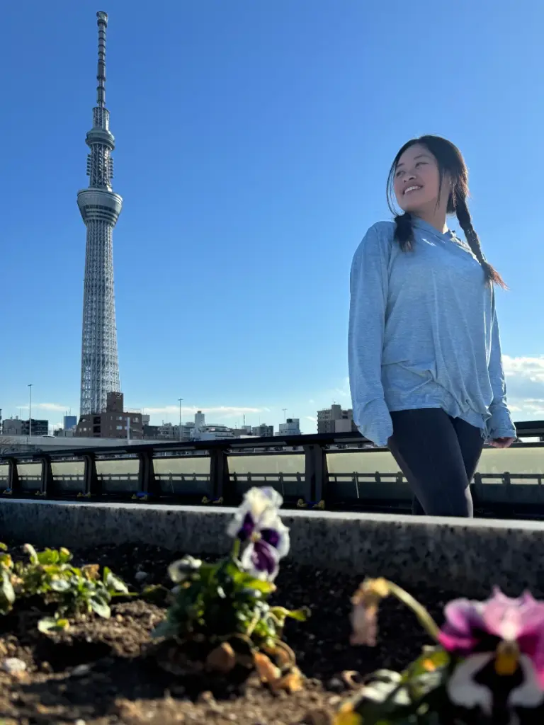 Zoe smiles while taking a break from a morning run along the Sumida River Terrace in Tokyo. The towering Tokyo Skytree stands tall against a clear blue sky in the background.