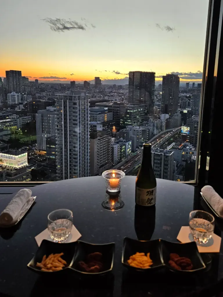 A bottle of sake, two glasses, and small snack bowls on a table next to a large window showing a golden sunset over the expansive Tokyo city skyline.