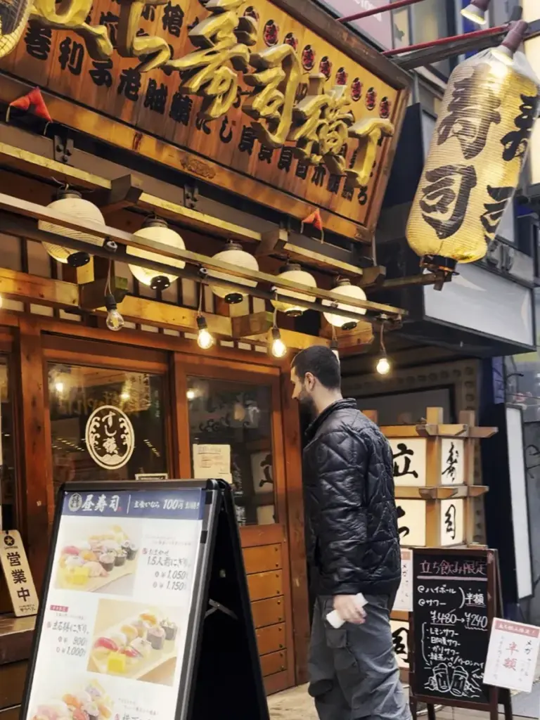 Tim walking into the wooden entrance of Tachi Sushi Yokocho, a popular standing sushi restaurant in Tokyo glowing with paper lanterns.