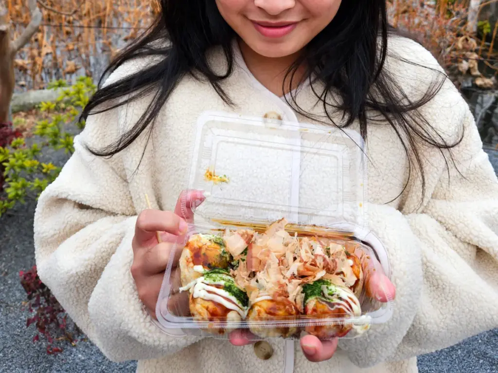 Zoe, wearing a cream-colored fuzzy jacket, smiling and holding a clear plastic container filled with fresh takoyaki (octopus balls) in Ueno Park. The takoyaki is topped with bonito flakes, kewpie mayo, and aonori (seaweed flakes).