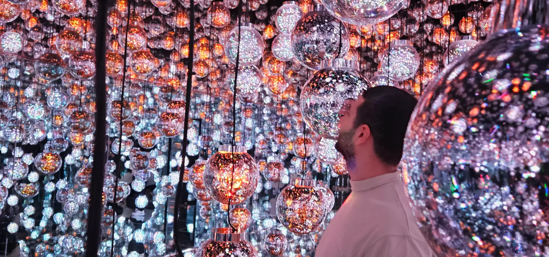 Tim looking up at hundreds of glowing, hanging glass spheres inside the mirrored Bubble Universe exhibit at teamLab Borderless.