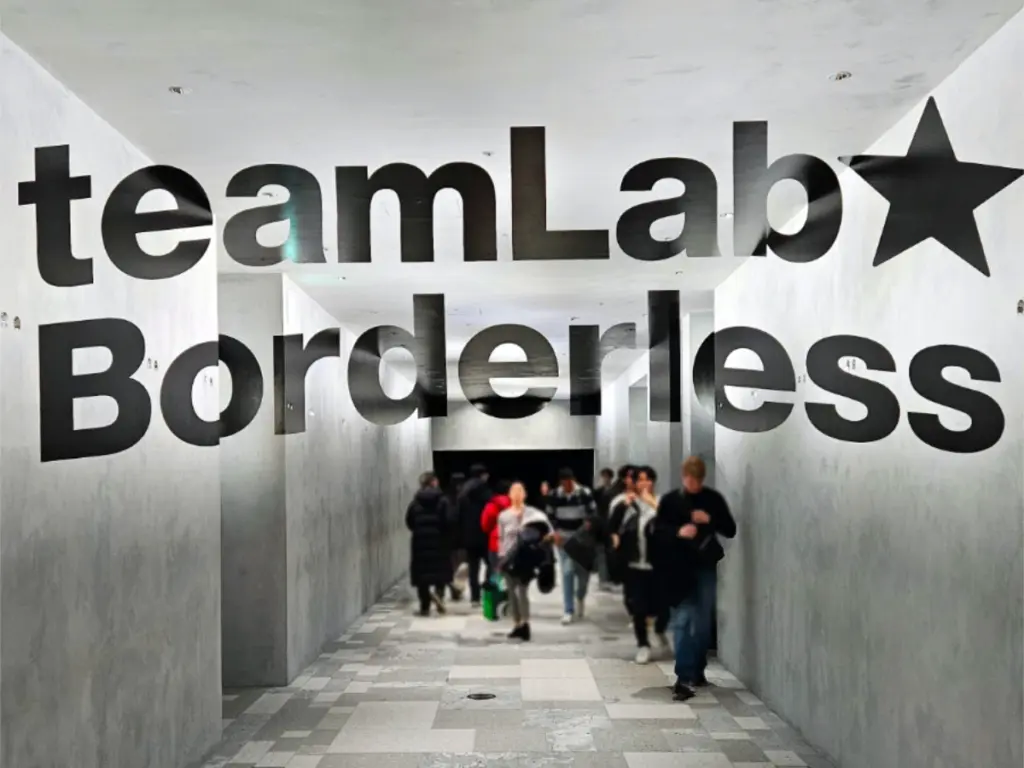 Large black lettering spelling out teamLab Borderless on a minimalist concrete wall at the entrance of the digital art museum in Tokyo, with visitors walking down the illuminated hallway.