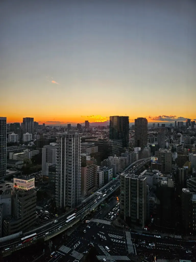 An aerial view of the Tokyo city skyline at sunset, with a vibrant orange horizon, tall skyscrapers, and a busy illuminated highway winding through the buildings below.