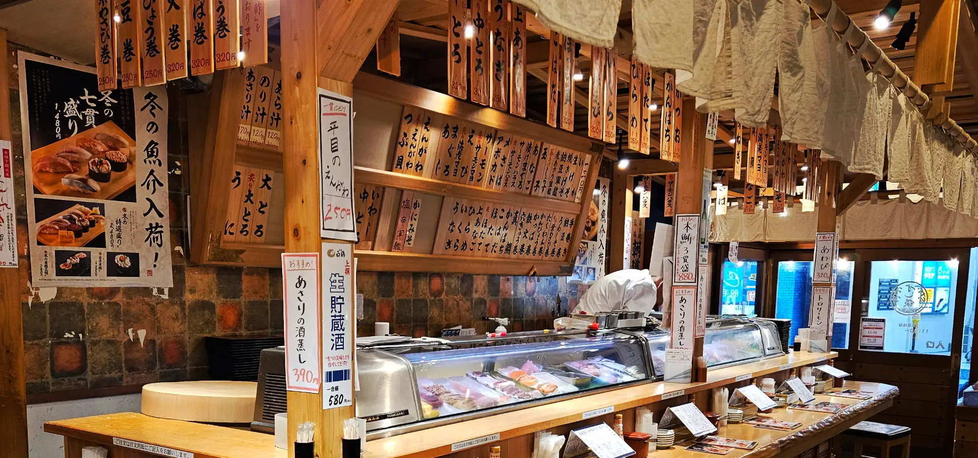 The warm, wooden interior of a traditional standing sushi restaurant in Tokyo, featuring a long dining counter, glass display cases filled with fresh fish, and hanging wooden menu boards with Japanese text.