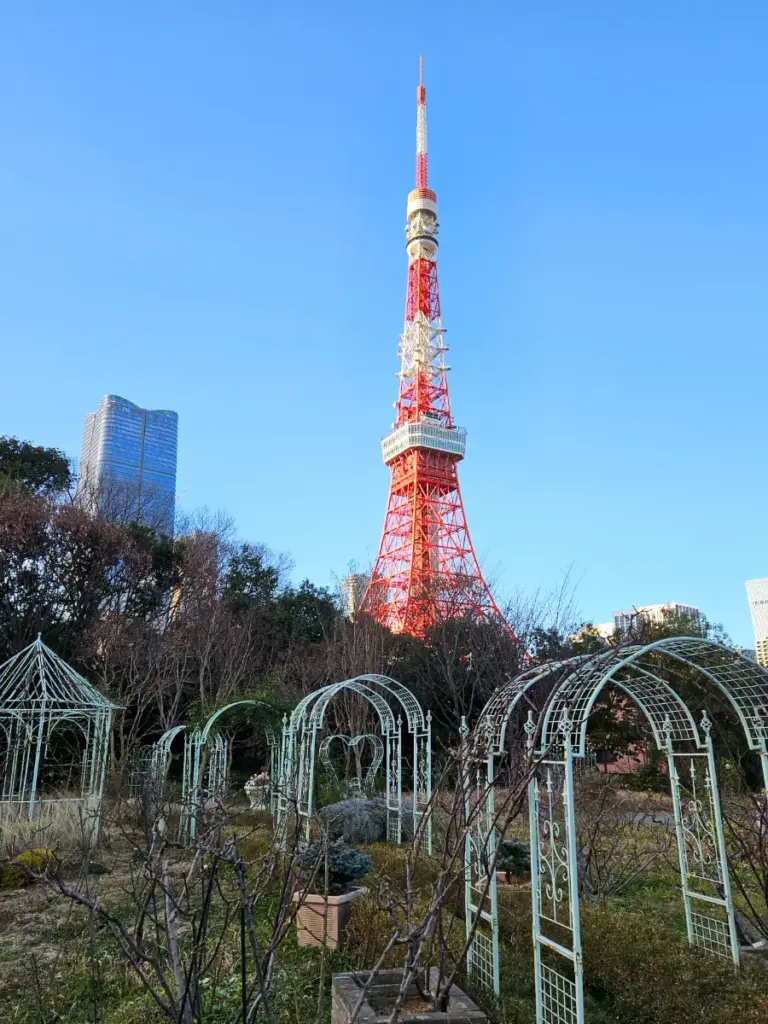 A daytime view of the Tokyo Tower rising above the winter trees, taken from a garden in Shiba Park with decorative green metal arches framing the pathway.