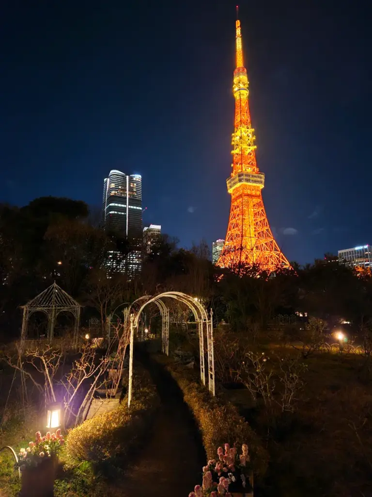 A nighttime view looking up at the glowing orange Tokyo Tower from a dark, peaceful garden pathway in Shiba Park, framed by illuminated white garden arches and distant city lights.