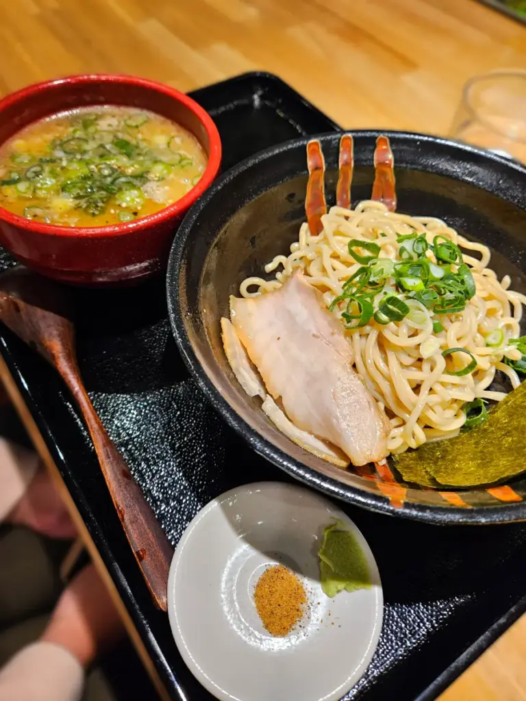 A black serving tray holding a bowl of thick tsukemen ramen noodles topped with sliced pork and green onions, sitting next to a red bowl of rich, savory dipping broth.