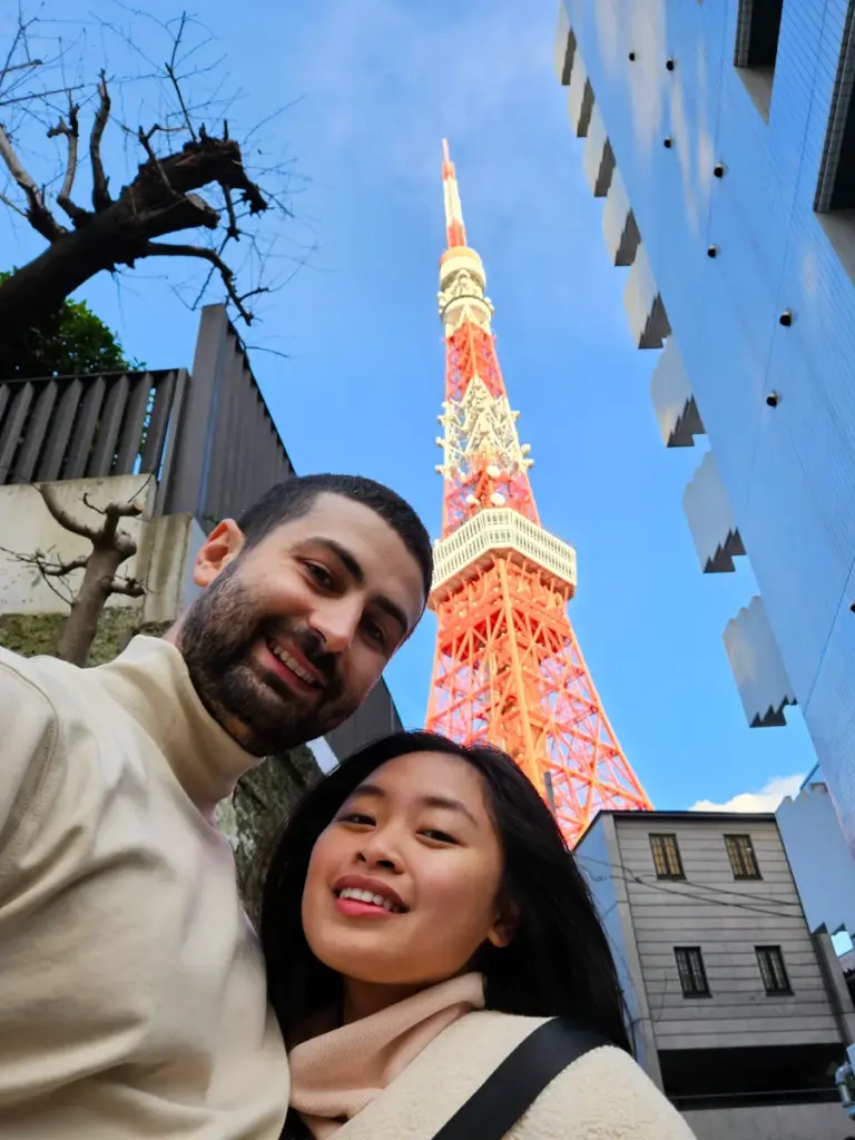 Zoe and Tim taking a selfie on a city street with the massive Tokyo Tower towering directly behind them against a bright blue sky.