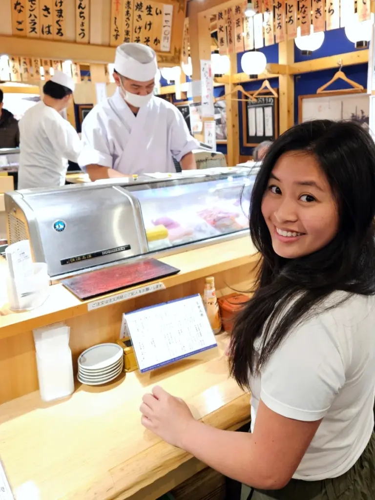 Zoe smiling brightly at the camera while standing at the wooden counter of a local Tokyo sushi restaurant, with a menu in front of her and a sushi chef working behind the glass display case.