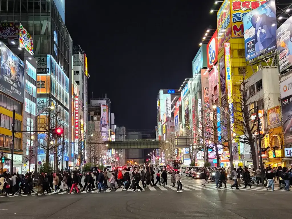 A large crowd of people crossing the street at night in Akihabara, Tokyo, surrounded by brightly lit neon signs and anime billboards.
