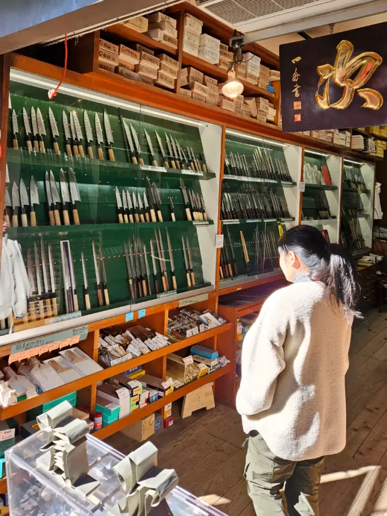 A woman looking at a large wall display of traditional Japanese chef knives inside a wooden shop in Tokyo.