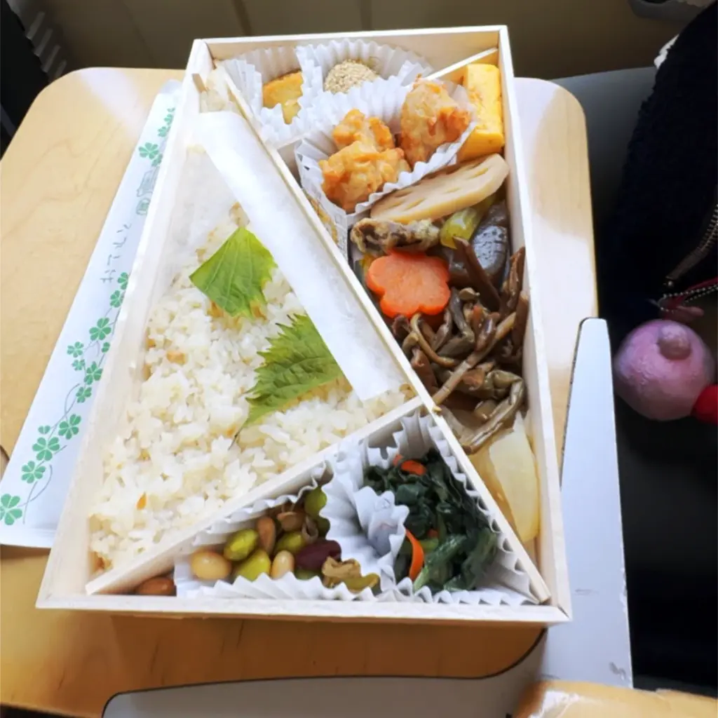 A traditional Japanese ekiben lunch box on a tray table, filled with rice, vegetables, and small side dishes.