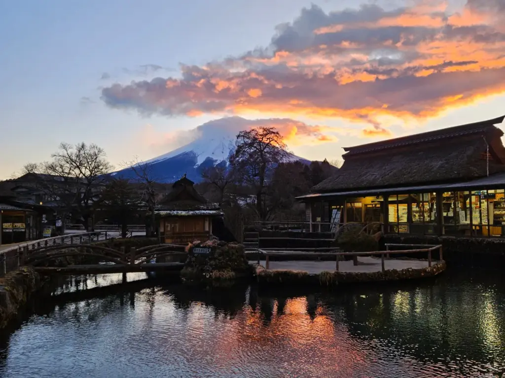 A stunning, peaceful view of the snow-capped peak of Mount Fuji at sunset, framed by a traditional thatched-roof building, a wooden bridge over a calm pond, and colorful orange and purple twilight clouds.