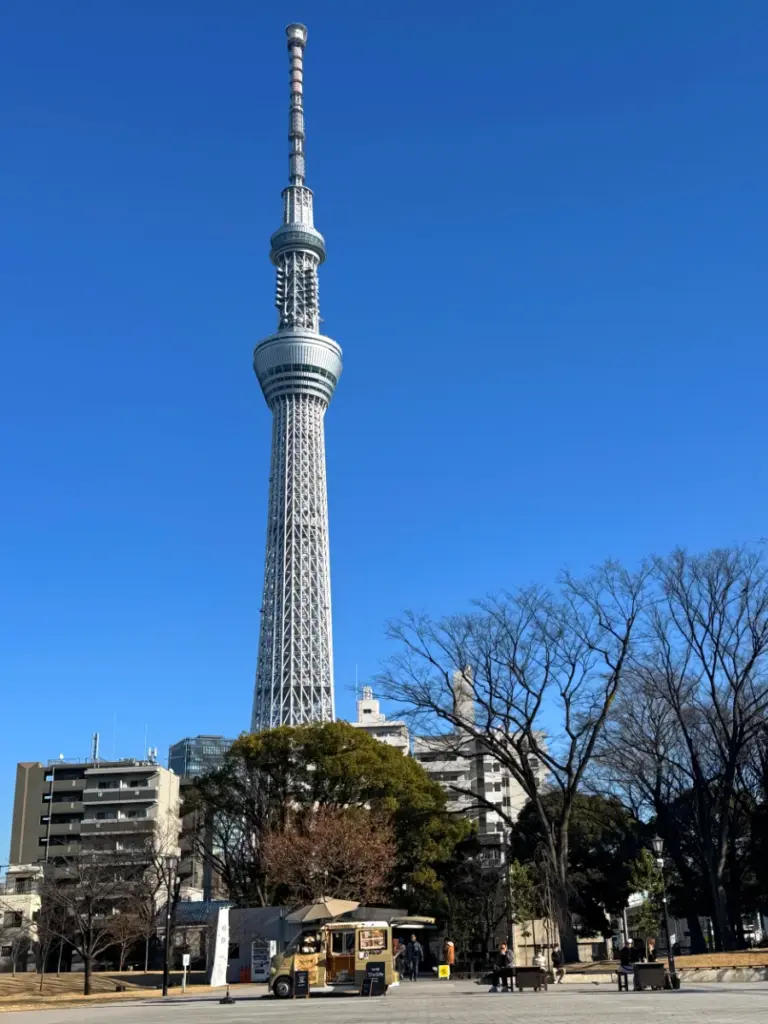 A daytime view of the towering Tokyo Skytree from Sumida Park Breeze Plaza, with a small vintage coffee truck parked in the foreground.