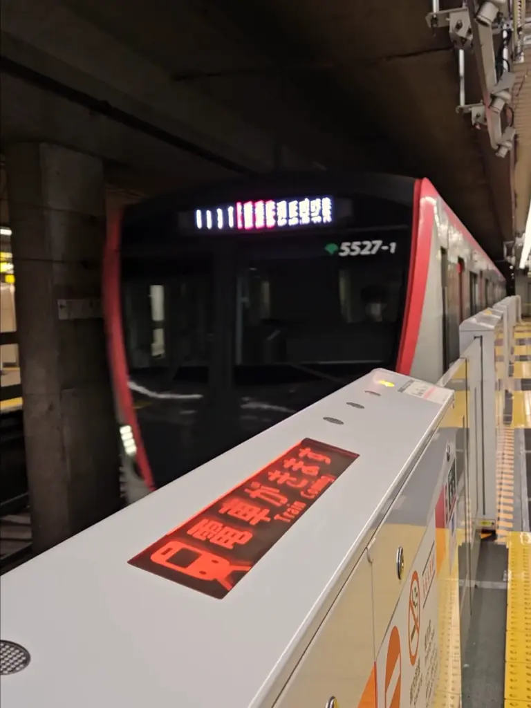 A red and white subway train arriving at an underground station platform in Tokyo, with a glowing red "Train Coming" warning sign on the safety barrier in the foreground.