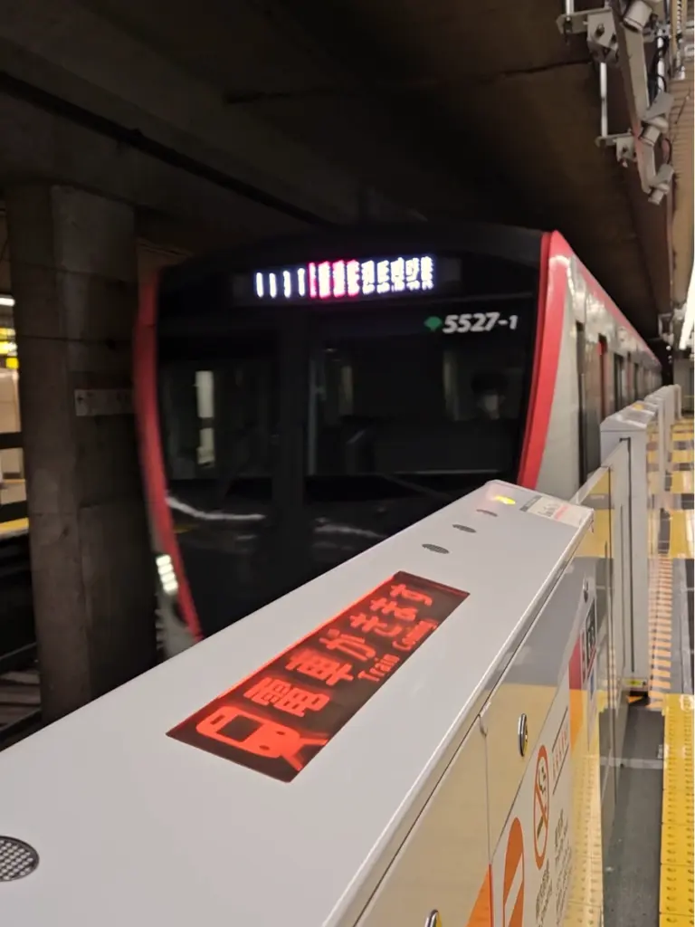A red and black Tokyo subway train pulling into an underground station, viewed from behind a safety barrier with a glowing red sign indicating the train is arriving.