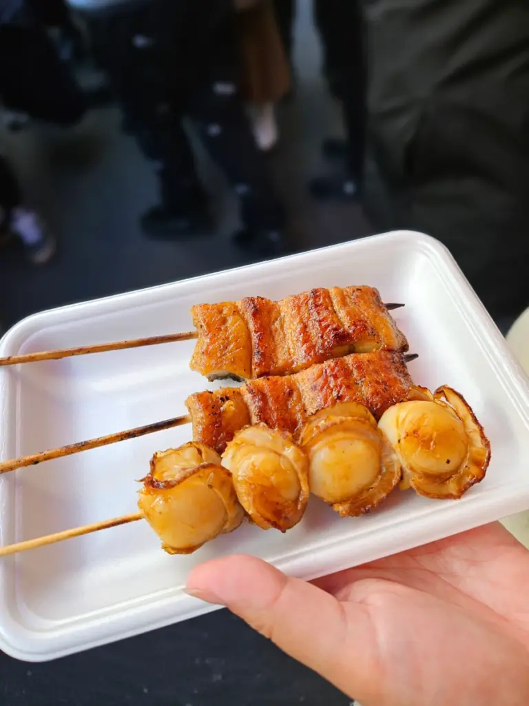 A hand holding a white tray with freshly grilled scallop and eel skewers from a street food vendor at the Tsukiji Outer Market in Tokyo.