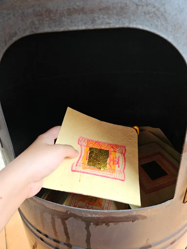 A hand placing traditional ghost money with a gold foil square into a burner during the Dragon Boat Festival.