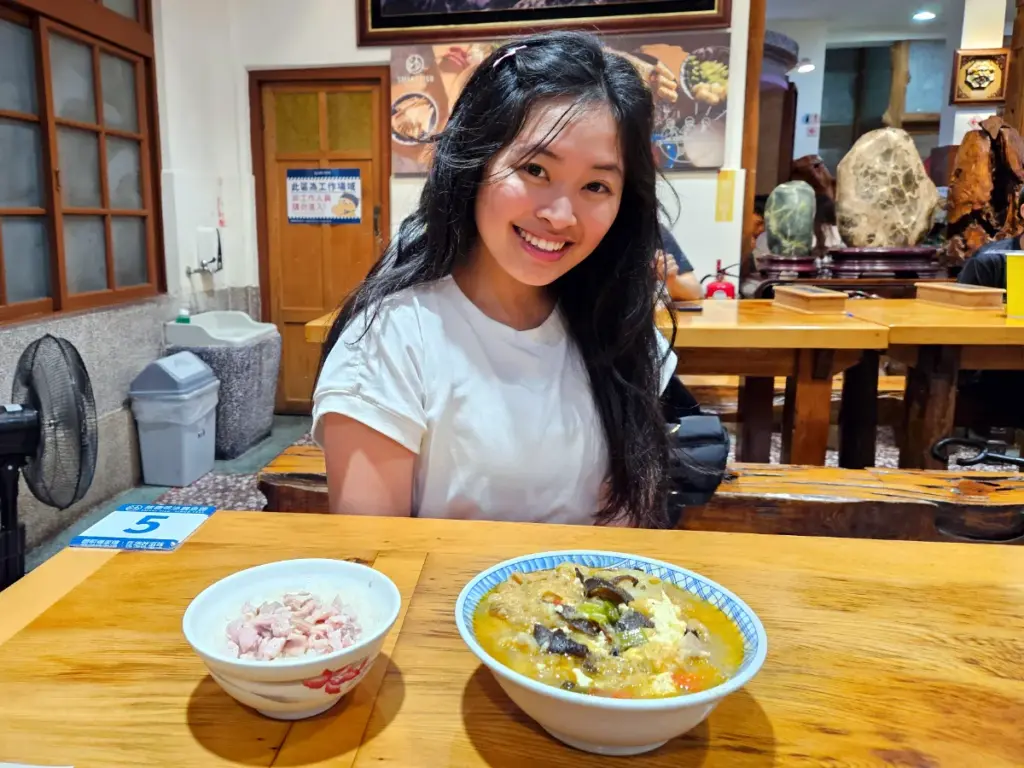 Zoe sitting at a wooden table with a bowl of Chiayi's famous fish head soup and local turkey rice.