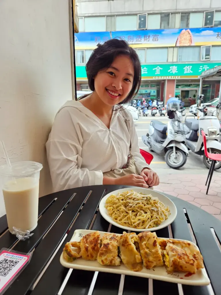 Zoe smiling at an outdoor table with a traditional local Taiwanese breakfast of dan bing, noodles, and iced soy milk.