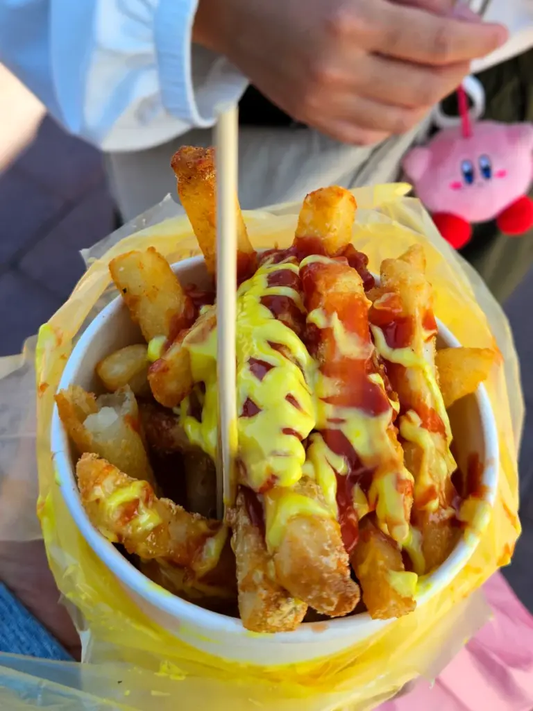 A cup of crispy french fries heavily drizzled with yellow mustard and red ketchup from a local festival food stall.