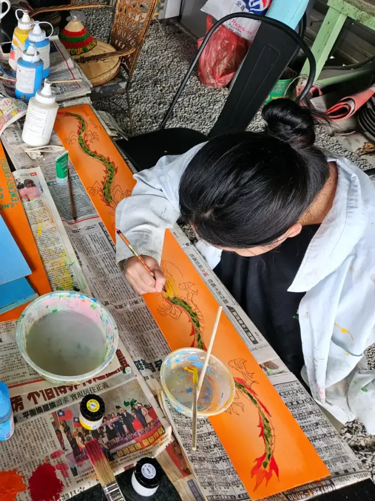 Zoe painting a colorful dragon on a wooden board over a newspaper-covered table during a rainy day at the tea shop.