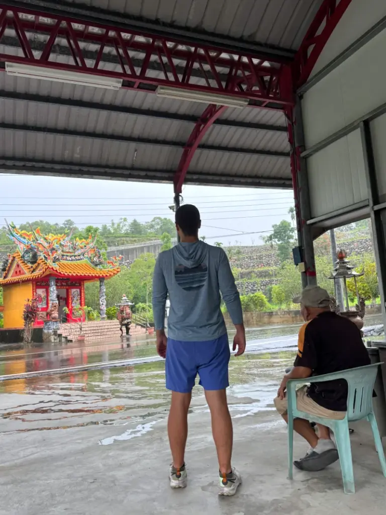 Tim standing and Steve sitting under a shelter, watching the heavy rain fall near a small temple during our Taiwan farm stay.