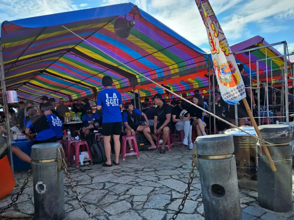 Festival-goers taking a break from the intense heat, sitting on red plastic stools under a brightly colored striped tent at the harbor.
