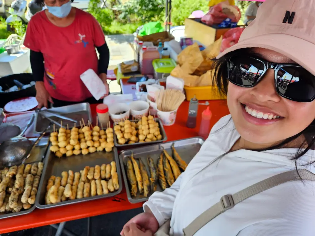 Zoe smiling in front of a bustling Taiwanese food stall selling skewers and fried seafood.
