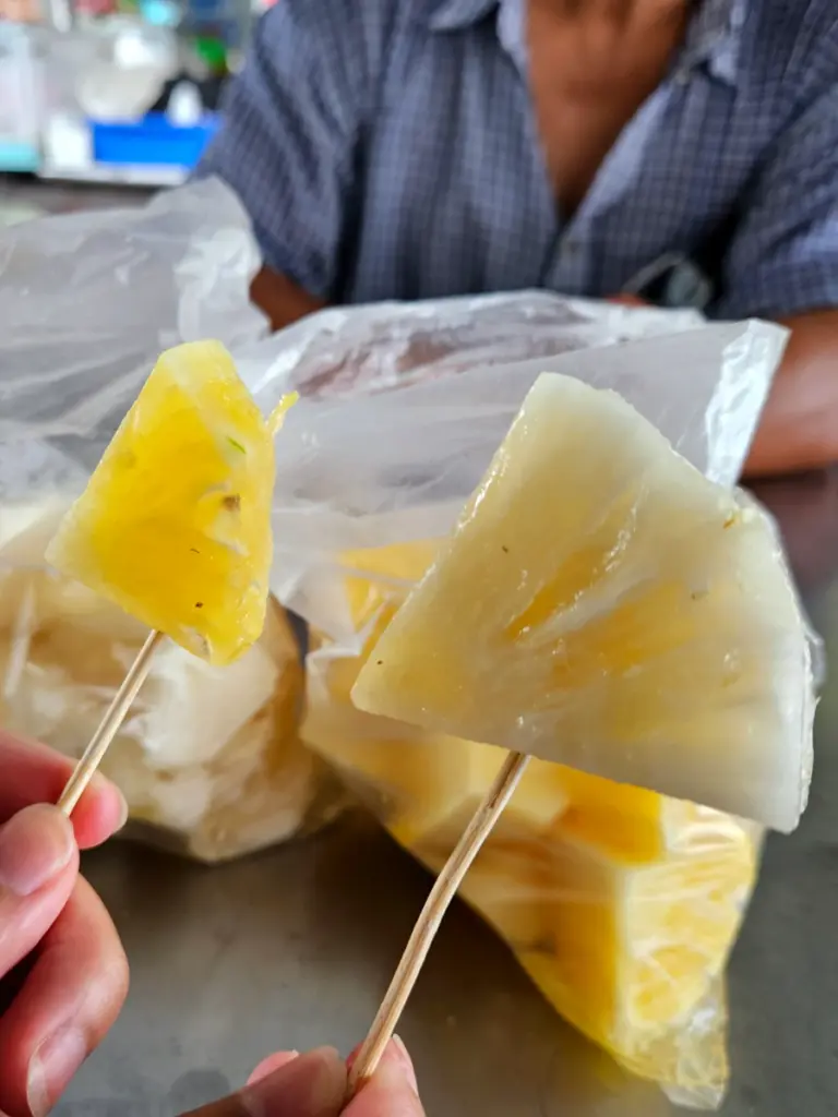 A hand holding two skewers comparing a regular golden pineapple to Taiwan's unique, creamy white milk pineapple.