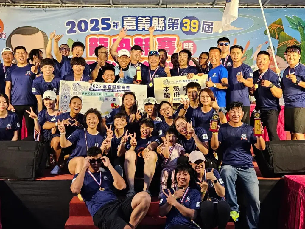 The winning dragon boat racing team celebrating enthusiastically on stage with a giant prize check and trophies.