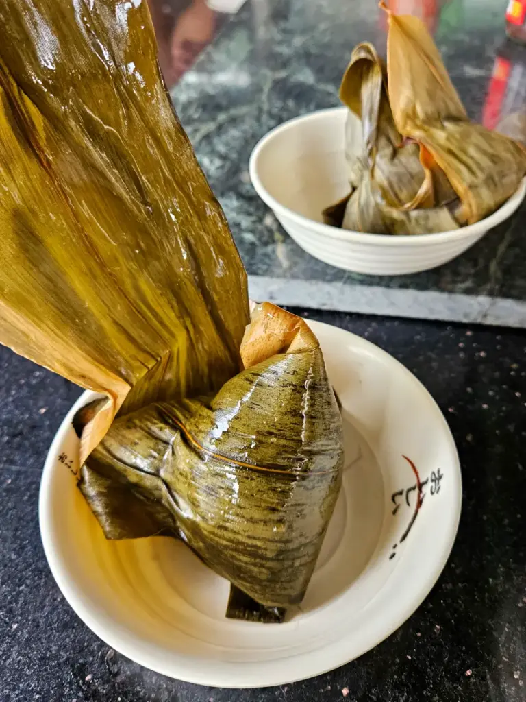 Traditional Taiwanese zongzi wrapped tightly in green bamboo leaves, resting in small white bowls.