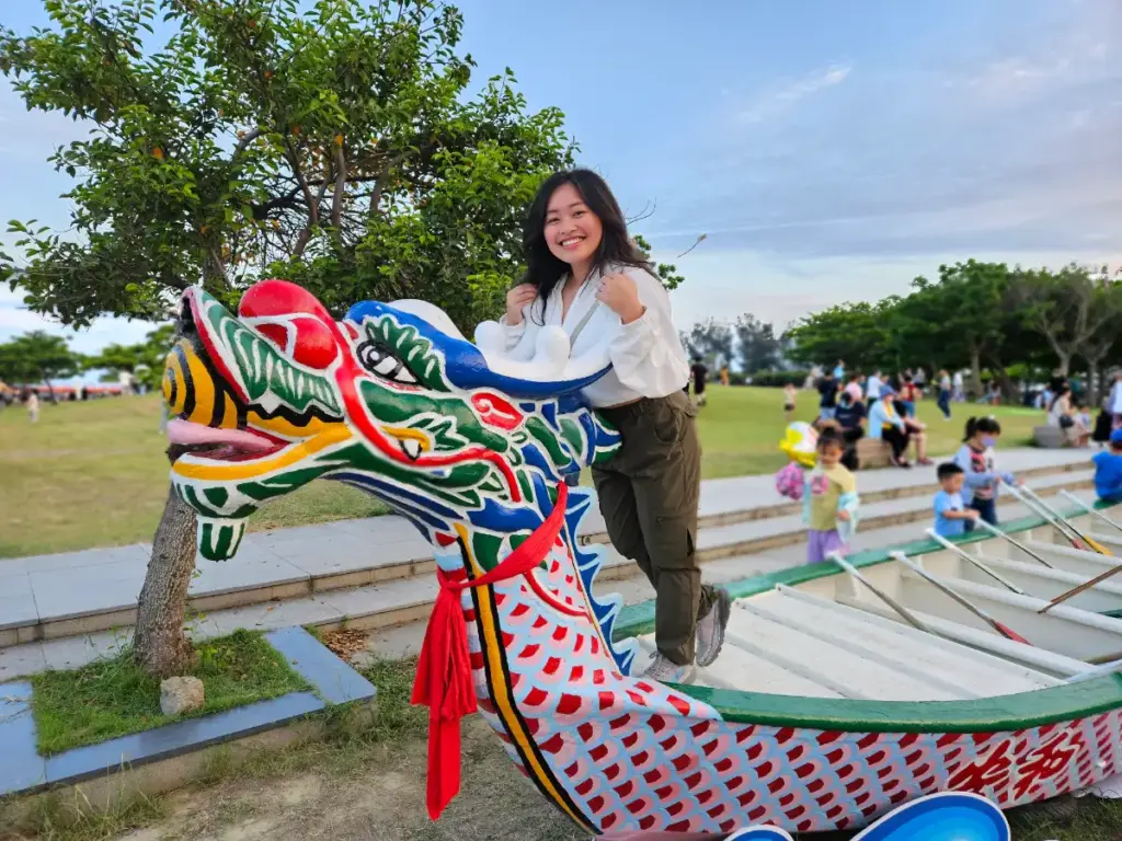 Zoe smiling while posing on a colorful, decorative dragon boat on the grass at the Dongshi Fisherman's Wharf.