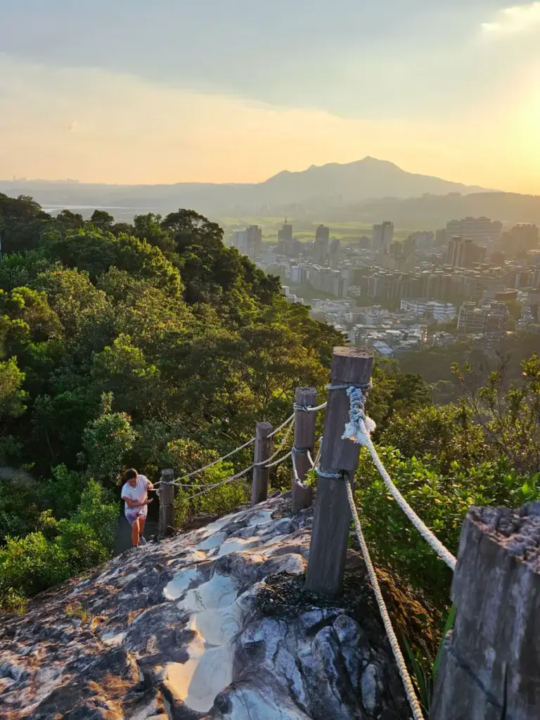 Zoe climbing a steep rock ridge trail lined with wooden rope posts overlooking the sprawling city basin at sunset.