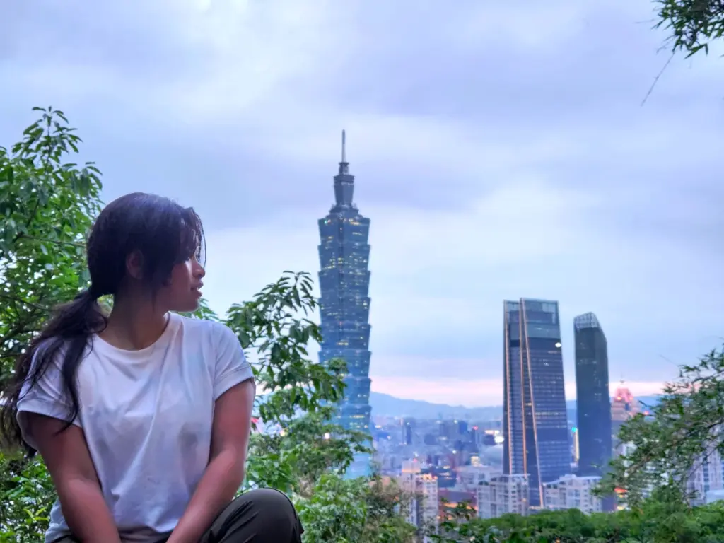 Zoe sitting on an overlook at Elephant Mountain at dusk, looking out over the illuminated Taipei 101 skyline.