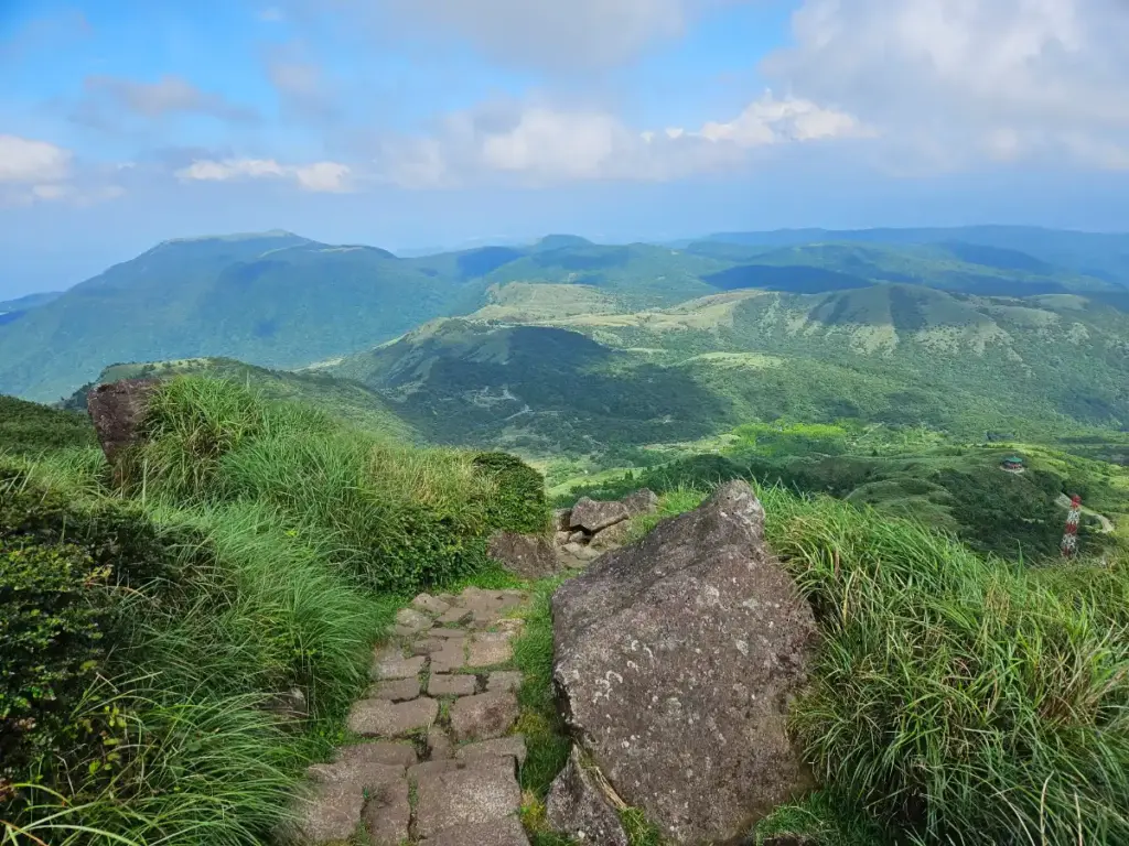 A stone-paved hiking path winding through lush green grass towards the rolling volcanic peaks of Qixing Mountain.