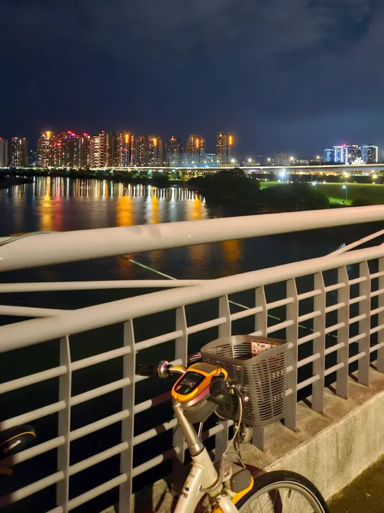 A rented yellow YouBike parked along a pedestrian bridge railing, overlooking a scenic Taipei riverfront with city lights reflecting on the water at night.
