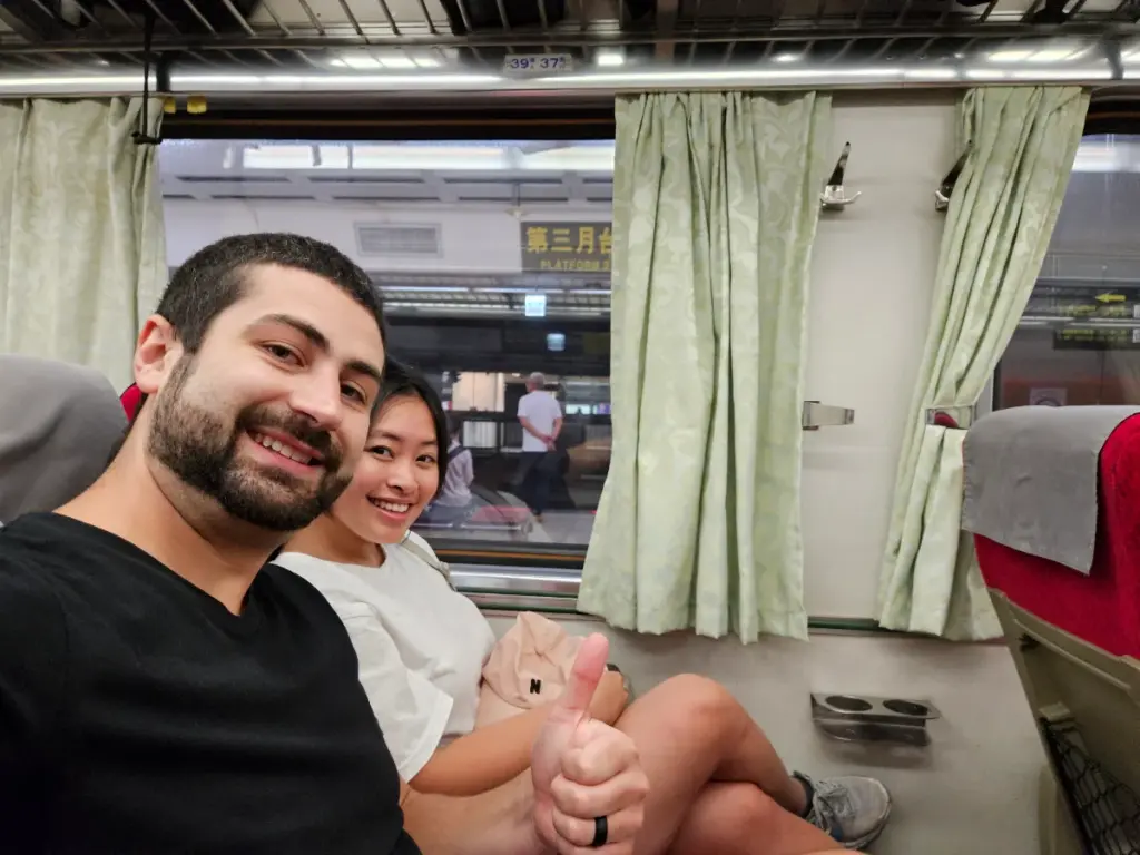 Tim and Zoe smiling and giving a thumbs-up while sitting next to a window inside a comfortable Taiwan Railway Administration (TRA) train carriage.