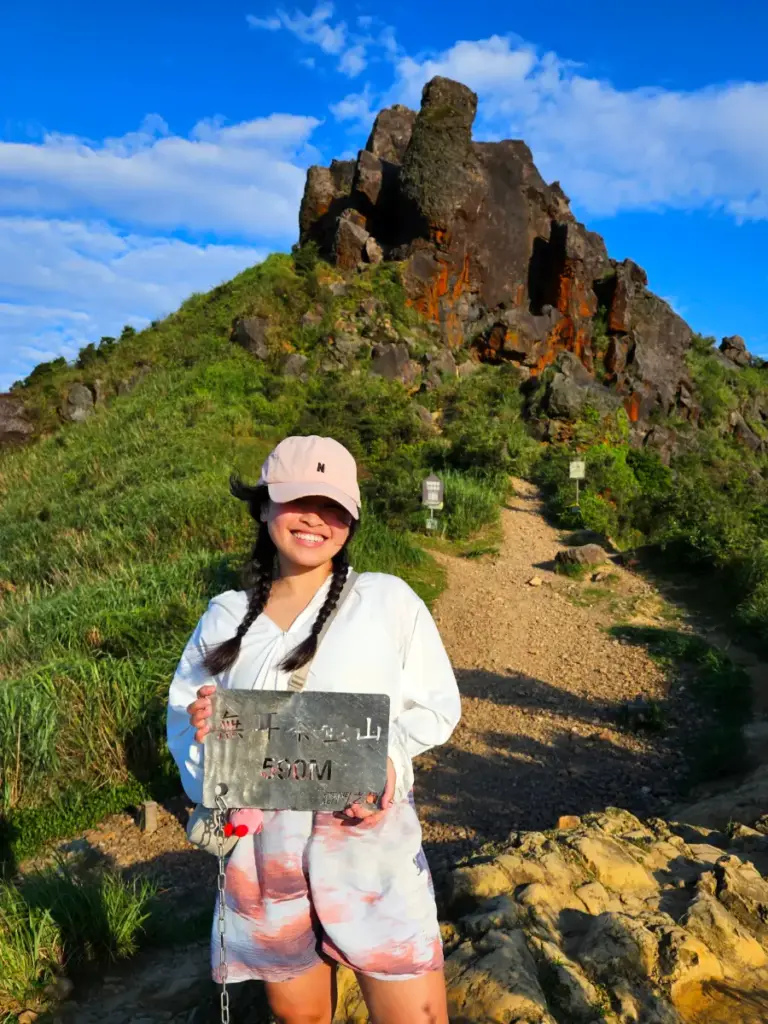 Zoe smiling and holding up the 580-meter summit sign in front of the rocky peak of Teapot Mountain.