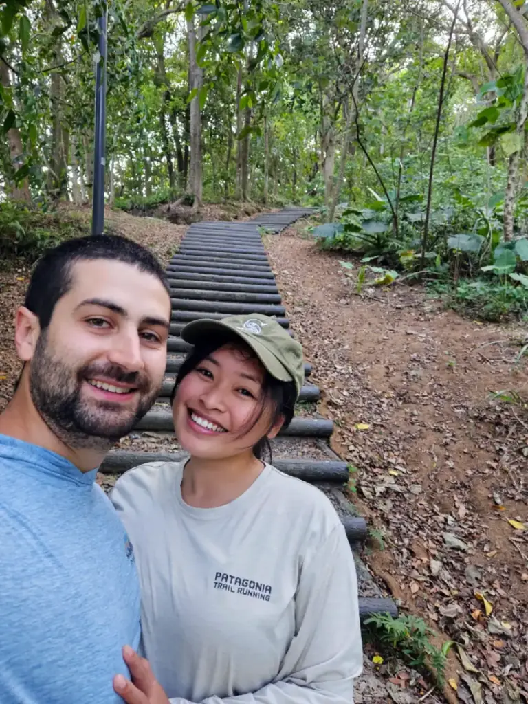 Tim and Zoe smiling together on a wooden log staircase trail surrounded by a dense forest canopy.